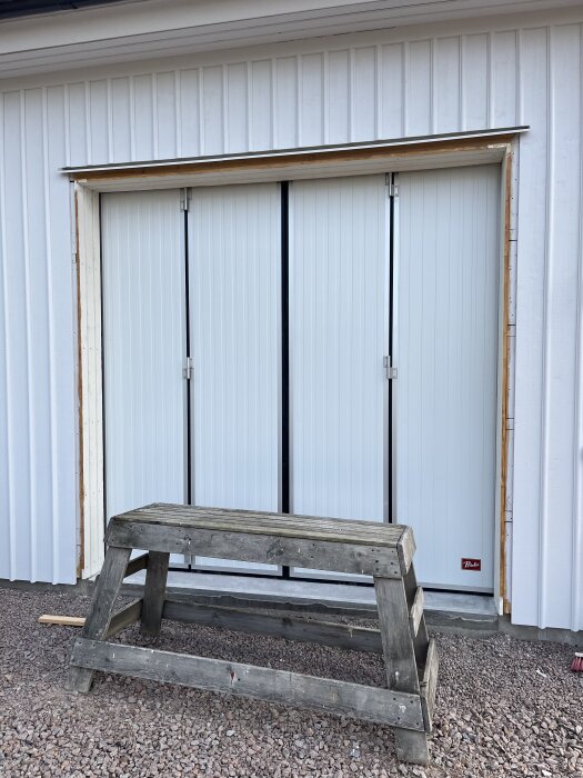 Dubbelportar målade i vitt med omgivande portinklädnad av trä på en vit fasad, träbockar står framför.