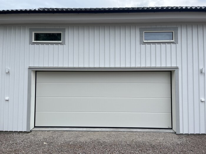 Vit garageport monterad på en byggnad med vita träpaneler och två små fönster ovanför.