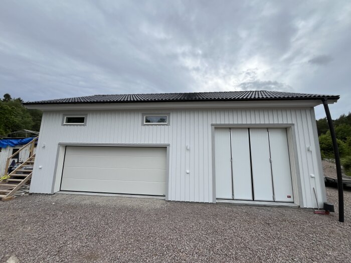 Vit byggnad med två portar och foder monterade, omgiven av grus och träd.