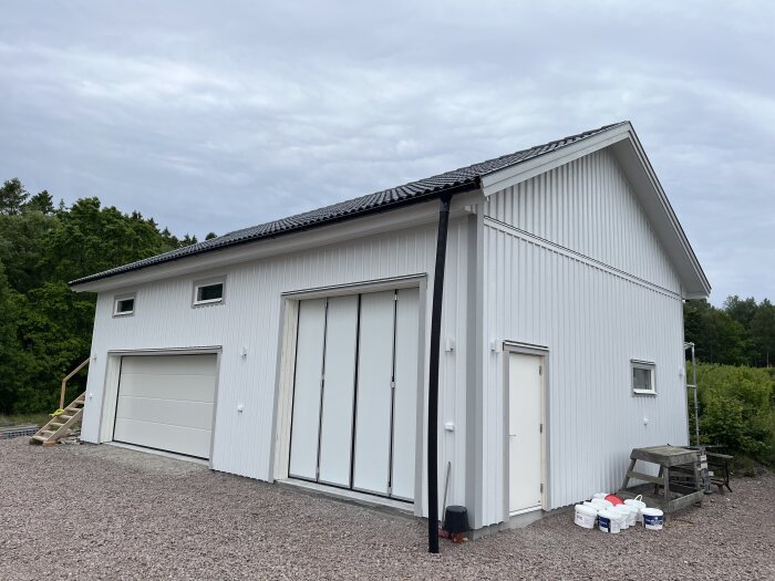 Vit byggnad med garageportar och träpanel. Vinklad takfot och omgivande grusplan. Skog syns i bakgrunden.