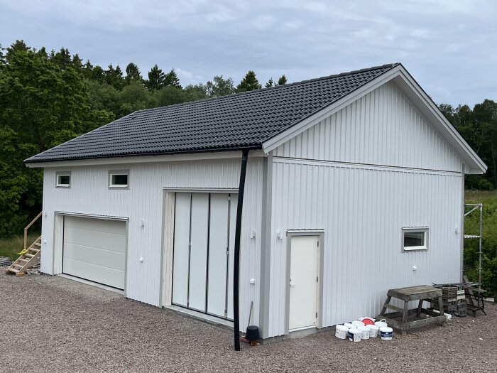 Vit byggnad med svart tak, flera portar och dörrar. Byggmaterial och ett bord syns framför byggnaden.