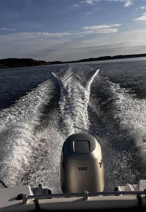 Båt i rörelse på vatten med synlig motor, lämnar ett skumspår efter sig, mot en bakgrund av öar och klar himmel.