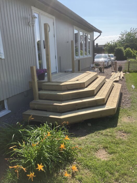 Trätrappa med flera steg vid ett hus, omgiven av grönska och blommor, räcke under uppbyggnad, solig dag.