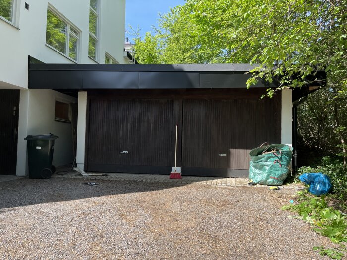 Grusad uppfart framför garage med två portar, soptunna till vänster, blå och grön säck med trädgårdsavfall till höger.