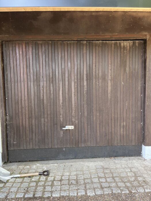 Träfärgad carportdörr med lodräta brädor, viss smuts, och en silversparke liggande framför.