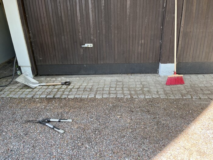 Carportdörrar i trä med smutsbeläggning, omgivna av en sop, spade och trädgårdsverktyg på marken.