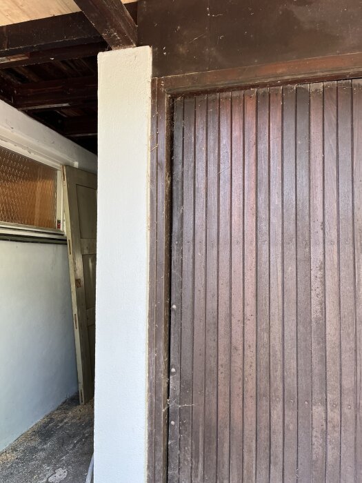 Sliten trädörr till en carport, täckt med spindelnät och smuts, omgiven av ljusa väggar och gammalt trä i ett hus från 1939.
