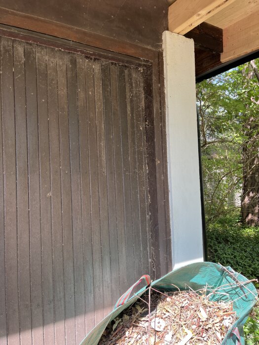 Trädörr till en gammal carport med smuts och spindelnät, samt en grön säck fylld med löv och kvistar.