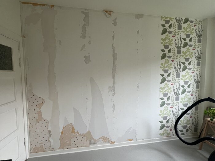Vägg med borttagen tapet, lämnar fläckar av gammal tapetlager och blottlägger körsbärsmönstrad tapet i hörnet. Ett lager med växttapet syns.