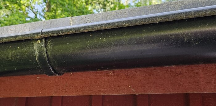 Svart stuprör och takränna utan repor, med röd träpanel i bakgrunden och gröna träd ovanför.