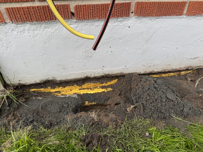 Marken vid husgrund med synlig gul dräneringsmatta och löst jord vid en vitmålad cementvägg och röda tegelstenar ovanför.