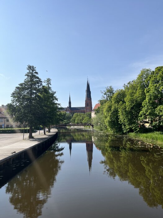 Flod med träd längs kanterna och en kyrka i bakgrunden som reflekteras i vattnet under en klarblå himmel.