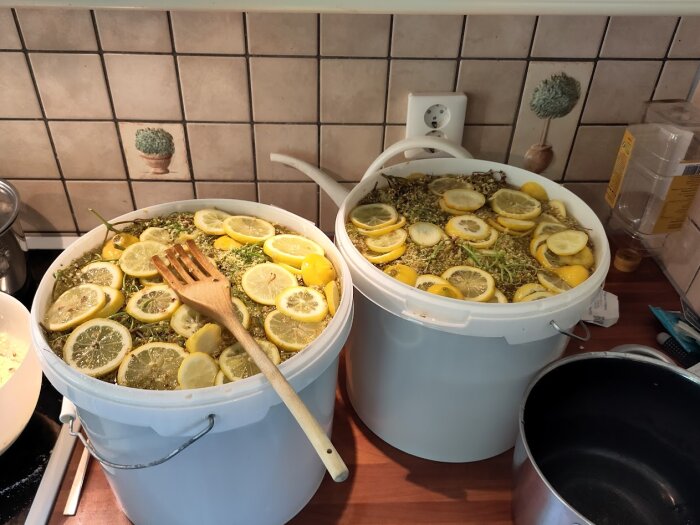 Två vita hinkar med citroner och fläderblommor står i ett kök. Träslev på kanten av en hink. Förberedelse av fläderblomssaft.