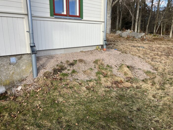Husvägg med grå stuprör, grusad sluttning och ojämn gräsmatta, orange avloppsdel och skog i bakgrunden.