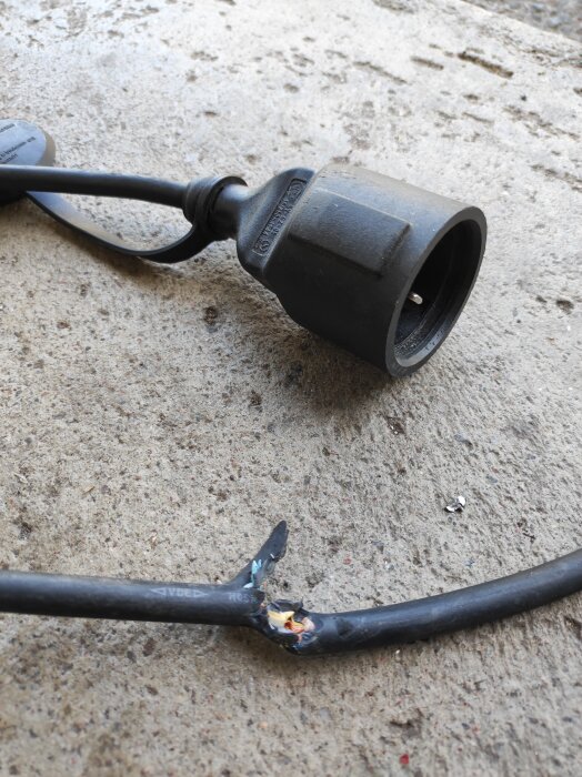 Svart elkabel med skadad isolering, exponerade ledningar och en kontakt placerad på ett betonggolv.