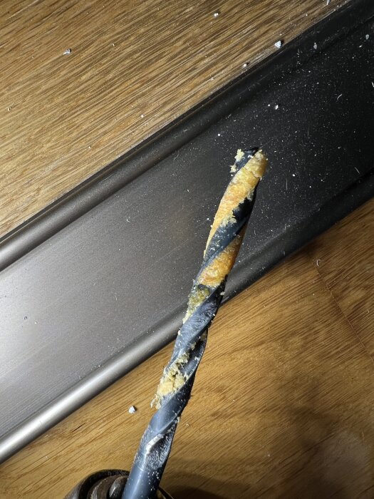 Borrspets med oljiga sågspån på träbord, visar resultatet av borrning i gammal väggregel, mindre synlig dammsamling runt omkring.