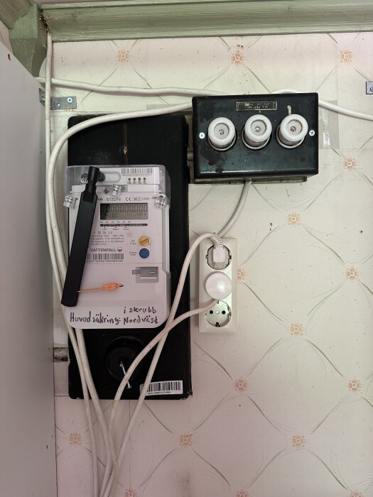 Bild på elskåp och huvudsäkring med flera kablar anslutna bredvid en vägg med blomtapet.