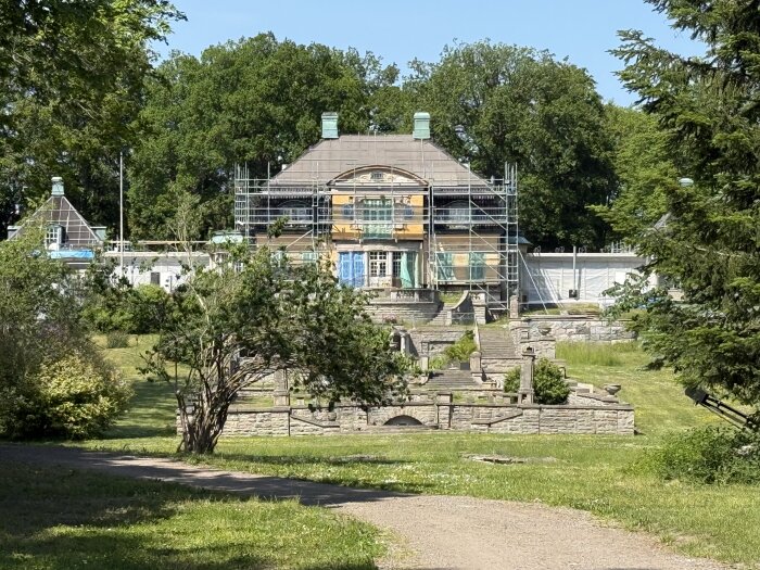 Hus under renovering med byggnadsställningar i trädgårdsmiljö, omgiven av träd och gräsmatta.