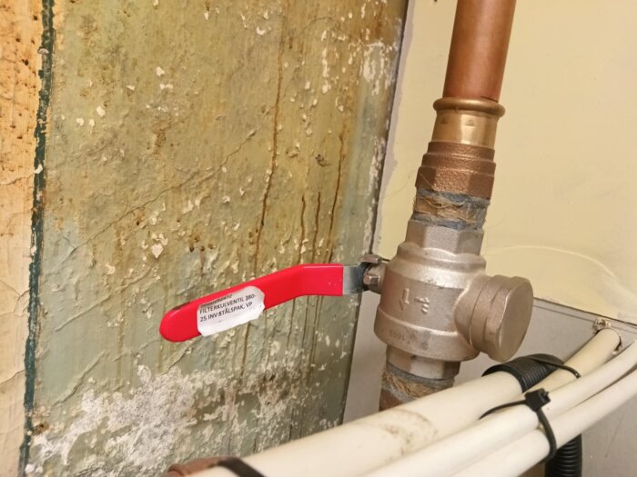 Filterkulventil med röd stålspak mot en gammal vägg, kopplad till kopparrör och plastslangar.