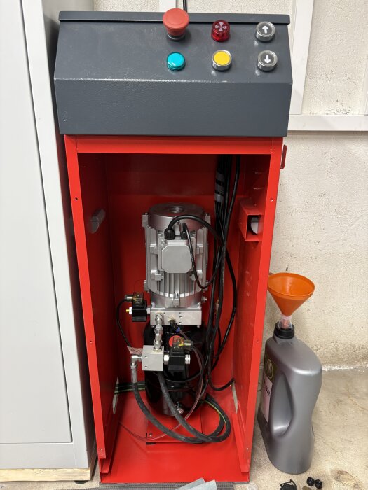 Röd elskåp med knappar på toppen, kablar och kopplingar inuti. Bredvid står en oljekanna med tratt. Framför lyftkonstruktion.