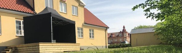 Scenbyggnad bakom gult ABF-hus med rött tak, omgiven av gräsmatta och träd, med en svart täckt del på scenen.