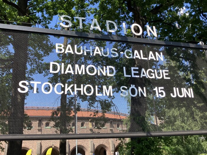 Skylt vid stadion annonserar "Bauhaus-galan Diamond League Stockholm sön 15 juni" under grönskande träd och blå himmel.