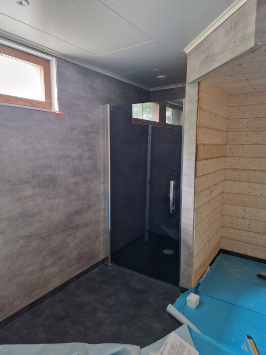 Badrum under renovering med nya väggpaneler och duschhörna, insvept i skyddsplast och byggmaterial på golvet.