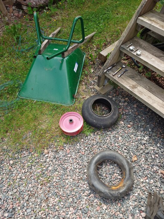 Omvänd skottkärra med demonterat hjul liggande bredvid, omgiven av grus och trätrappa med verktyg.