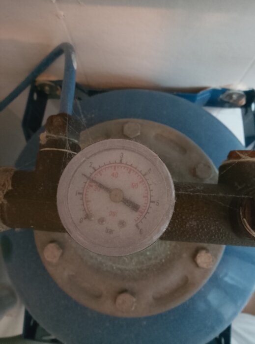 Manometer på en hydrofor visar trycknivåer i psi och bar, omgiven av damm och spindelväv, del av vattensystem för borrad brunn.