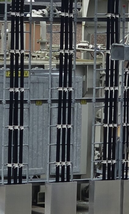 Svartisolatorer och kablar i elstation, hållna med vita band mot stödisolatorer, med ett grått hölje i bakgrunden.