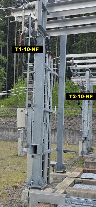 Två metallstrukturer märkta "T1-10-NF" och "T2-10-NF" i en utomhus miljö med kablar och elkomponenter.