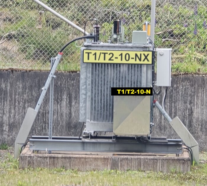 Transformator med märkning "T1/T2-10-NX" och "T1/T2-10-N", placerad på en fast bas, omgiven av vegetation och staket.