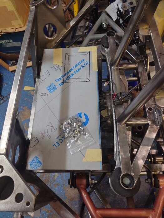 Metallram med kapad plåt och plastpåse med små skruvar, omgiven av arbetsyta med verktyg och delar.