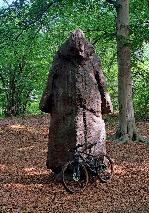 Stor stenstaty i en skog med en cykel lutad mot den, omgiven av gröna träd och brun fallna löv.