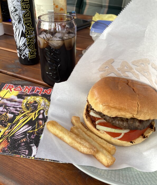 Hamburgare med pommes, dryck i Coca-Cola-glas och Iron Maiden-förpackning på trämöbel utomhus.