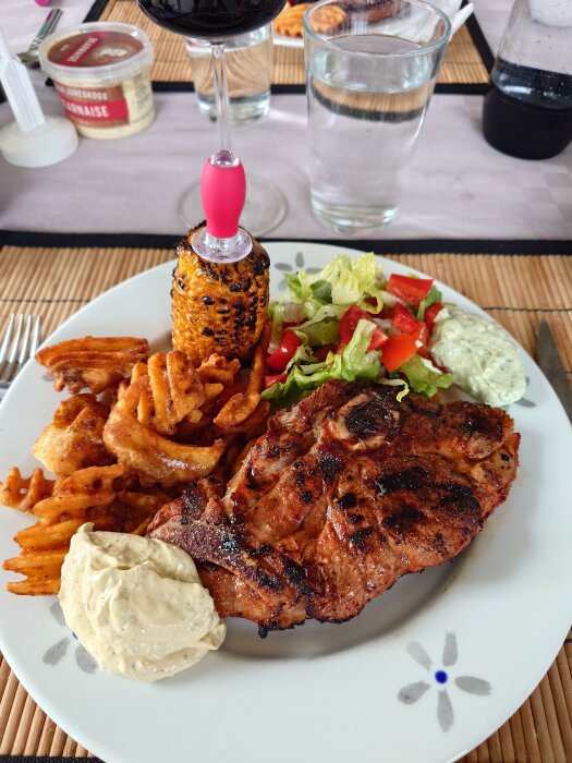 Tallrik med grillad flintastek, crisscuts, grillad majs, sallad och två såser. Vin- och vattenglas i bakgrunden.