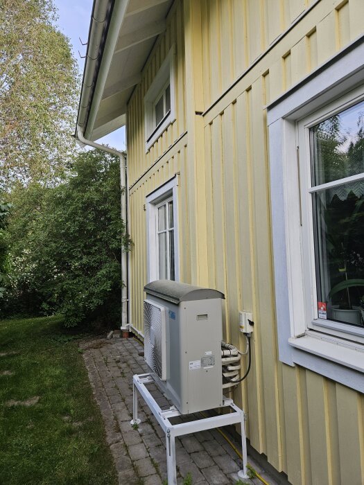 Utedel av en AC-enhet vid en gul husfasad med brädpanel och vitmålade fönster, omgiven av grönska.