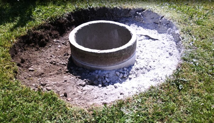 Cirkulär betongfördelningsbrunn i marken med tillhörande gräsyta och jord, omgiven av grus och cementdammsrester.