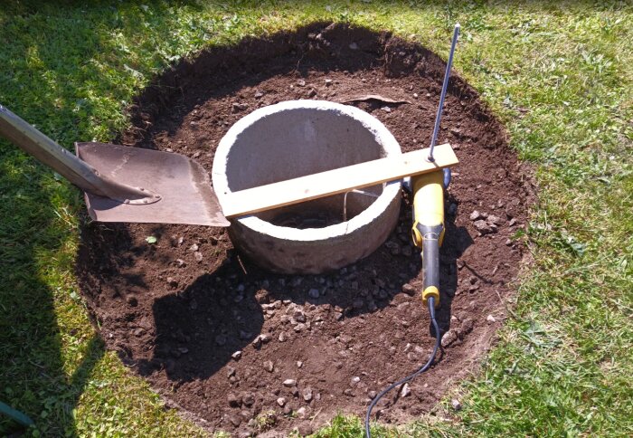 En fördelningsbrunn i trädgård, omgiven av jord och gräs, med en skyffel och en gul vinkelslip placerade vid kanten av brunnen.