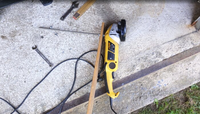 En gul vinkelslip ligger på betong tillsammans med verktyg, trämätsticka och kabel.