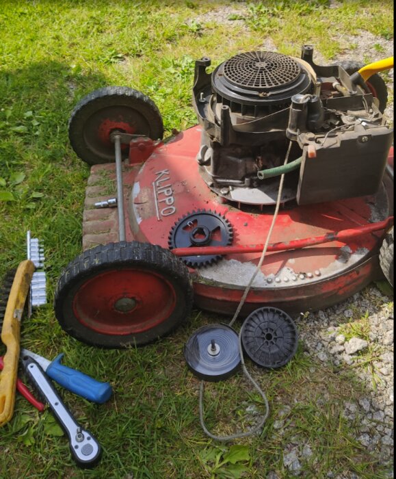 Röd gräsklippare med borttagna hjul och verktyg liggande på gräset, under reparation.