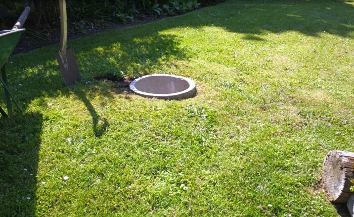Rund fördelningsbrunn i gräsmatta, omgiven av grönska. Skyffel och skottkärra syns vid kanten.