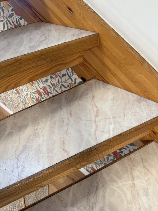 Trappa med trappsteg täckta av utskuren vinylmatta med trästruktur i kanten, placerad mot en tapetserad vägg med blomstermönster.