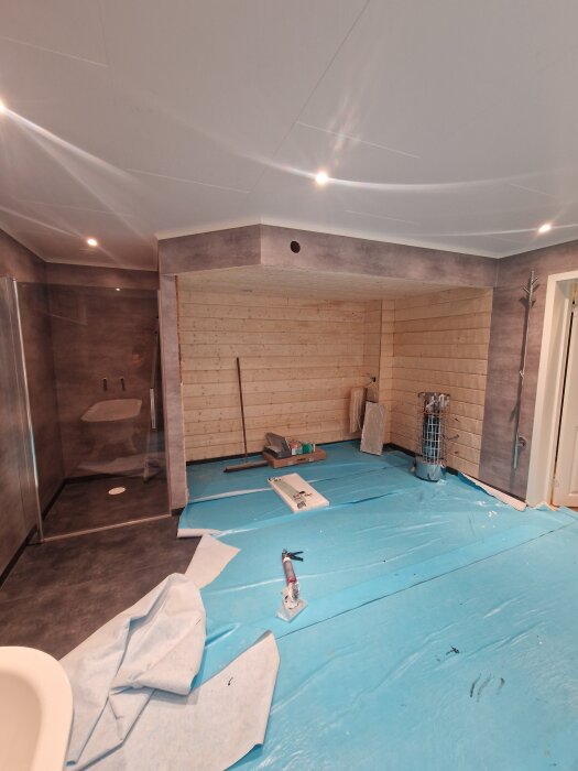 Rum under renovering med blått golvskydd, träpanelvägg, verktyg och material utspridda på golvet, del av badkar syns.