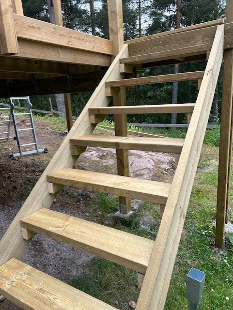 Trätrappa under konstruktion utomhus med plankor monterade på reglar; omgivning av skog och gräs.