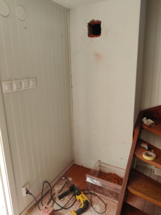 Vägg med hål för sotlucka under installation i fritidshus, med verktyg som slagborrmaskin och dammsugare på golvet.