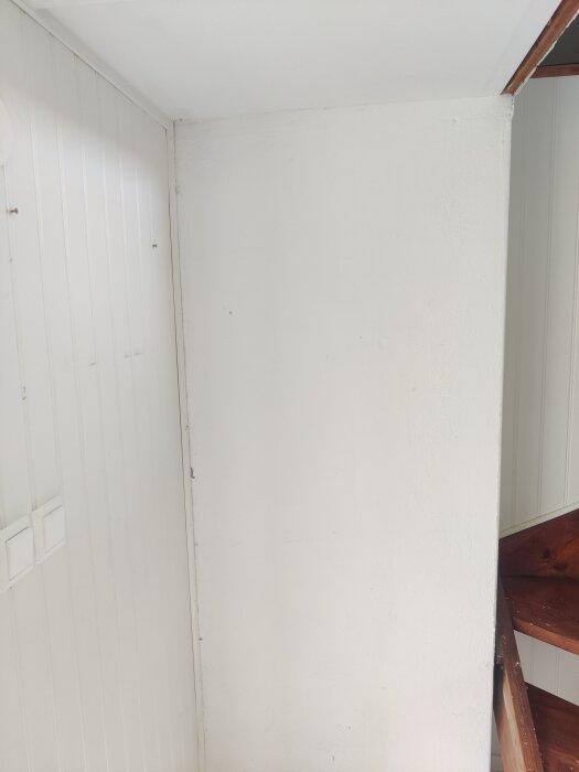 Vitt vägghörn i fritidshus, delvis synliga trappsteg av trä.