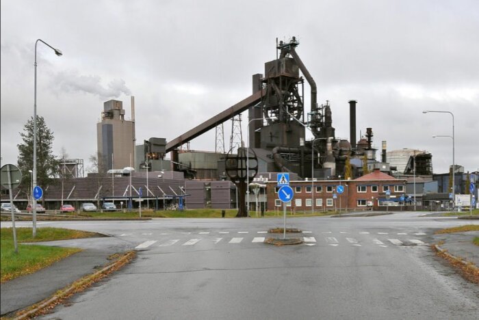 Industriell anläggning med skorstenar och utrustning, byggnader i förgrunden och ett övergångsställe framför, under en molnig himmel.