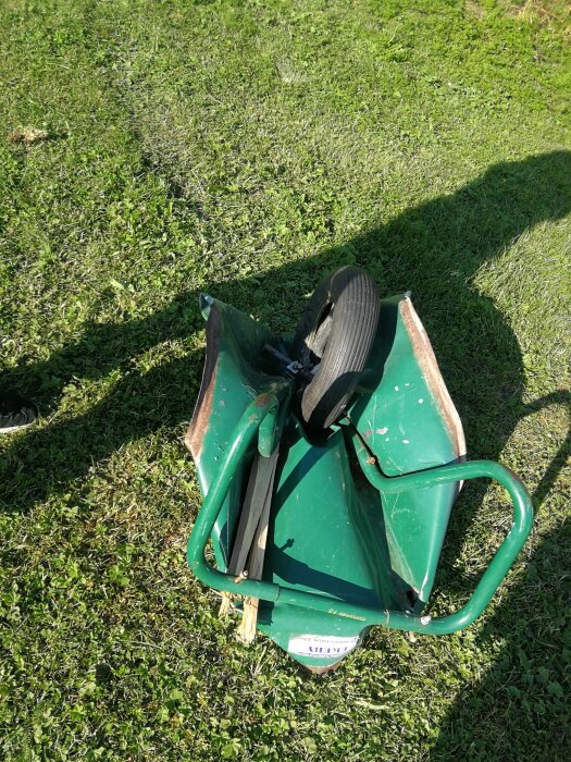 Grön skottkärra med trasigt handtag och defekt framhjul på gräsplätt.