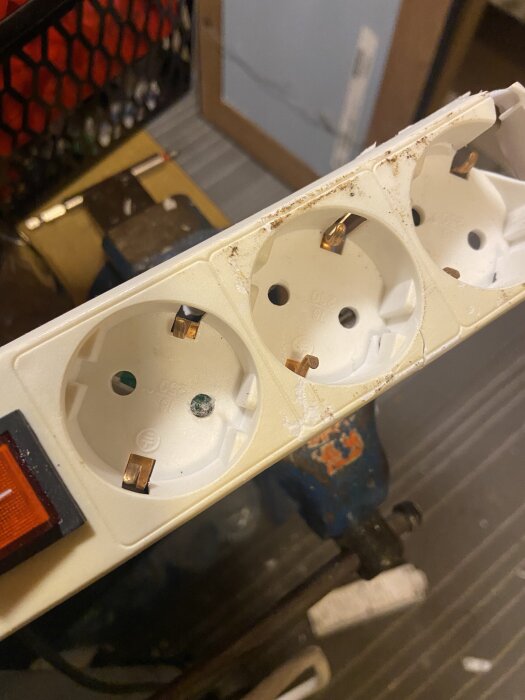 En vit grendosa med flera eluttag, varav några har synliga kopparbleck, visas i närbild. En röd strömbrytare syns på sidan.